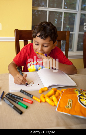 Eight-year-old Mexican-American boy snacks on junk food, Cheetos, while drawing at home with markers. Stock Photo