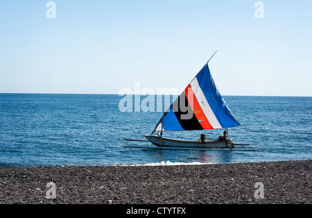 A traditional Balinese outrigger fishing boat, called a jukung, sails along the Amed, Bali coastline on a beautiful sunny day. Stock Photo