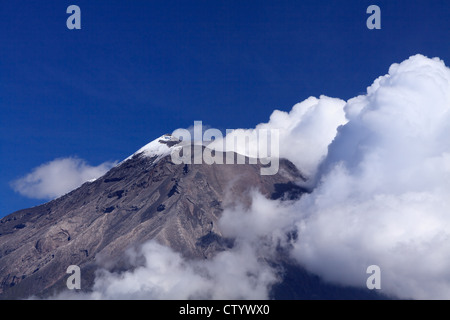 A rugged, snow-capped mountain peak rises sharply against a deep blue sky, partially enveloped by large, billowing white clouds. The mountain's slopes are steep and rocky, with visible striations and Stock Photo