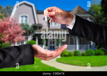 A hand giving a key to another hand. Both persons in suits and a house in the background. Stock Photo