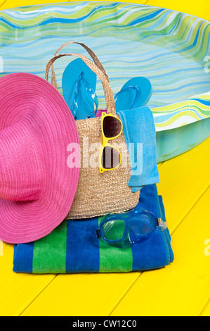 A straw beach bag full of accessories rests on a beach towel next to a children's plastic swimming pool.