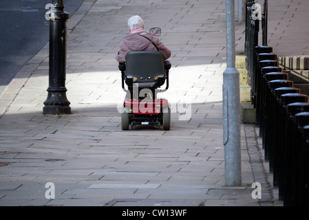 Elderly Woman ascending a steep rise in an electric Mobility Scooter  on the pavement in the UK Stock Photo