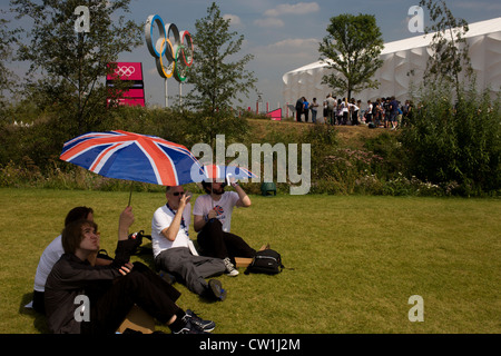 Brits enjoy a hot summer to watch live coverage from a large tv screen in the Olympic Park during the London 2012 Olympics. Under union jack umbrellas they sit on green grass located on a hilltop across from giant Olympic rings used as a background for spectators' photos. London’s Olympic Park, at just under a square mile, is the largest new park in the city for more than 100 years. The planting of 4,000 trees, 300,000 wetland plants and more than 150,000 perennial plants plus nectar-rich wildflower make for a colourful setting for the Games. (More captions in Description..) Stock Photo