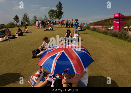 Brits enjoy a hot summer to watch live coverage from a large tv screen in the Olympic Park during the London 2012 Olympics. Under union jack umbrellas they sit on green grass located on a hilltop where giant Olympic rings act as a backdrop to spectators' family photos. This land was transformed to become a 2.5 Sq Km sporting complex, once industrial businesses and now the venue of eight venues including the main arena, Aquatics Centre and Velodrome plus the athletes' Olympic Village. After the Olympics, the park is to be known as Queen Elizabeth Olympic Park. (More captions in Description..) Stock Photo