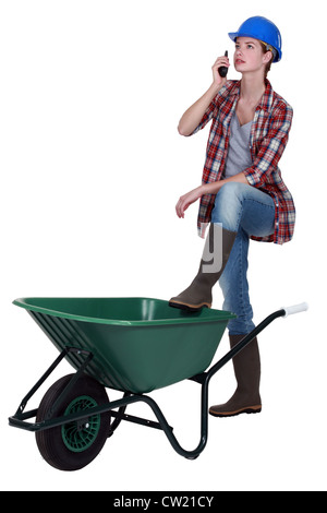 Tradeswoman speaking into a walkie-talkie and propping her foot up on a wheelbarrow Stock Photo