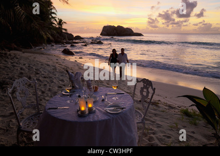 Romantic couple walking at sunset on beach with dinner table in foreground.Cousine Island.Seychelles Stock Photo