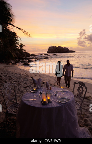 Romantic couple walking at sunset on beach with dinner table in foreground.Cousine Island.Seychelles Stock Photo