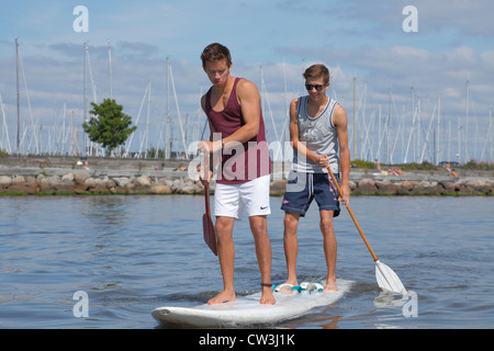 Two teenagers having fun stand up paddling on a windsurf board on a sunny summer day at the beach at Rungsted Harbour, Denmark. Paddleboarders Stock Photo