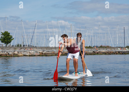 Two teenagers having fun stand up paddling on a windsurf board on a sunny summer day at the beach at Rungsted Harbour, Denmark. Stock Photo