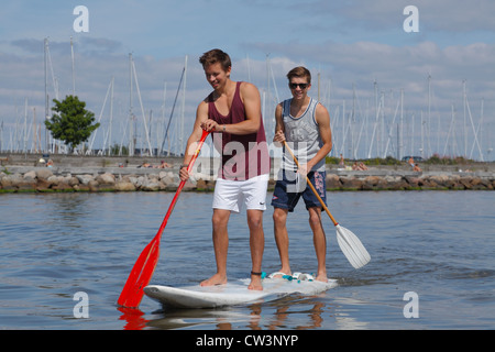 Two teenagers having fun stand up paddling on a windsurf board on a sunny summer day at the beach at Rungsted Harbour, Denmark. Stock Photo