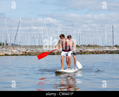 Two teenagers having fun stand up paddling on a windsurf board on a sunny summer day at the beach at Rungsted Harbour, Denmark. Paddleboarders Stock Photo