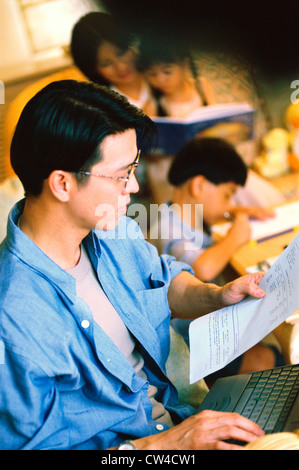 Side profile of a father working on a laptop with his family sitting behind him Stock Photo