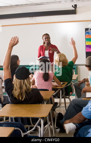 Cheerful female teacher holding book and students raising hands in the classroom Stock Photo