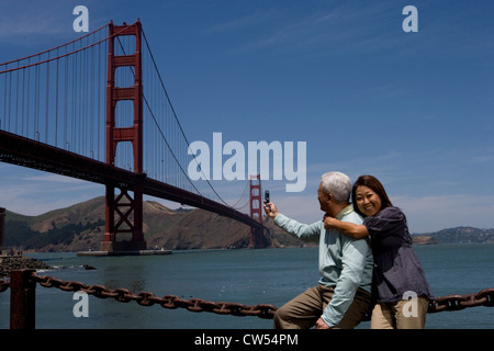 Mature man taking picture suspension bridge with mature woman embracing him from behind Golden Gate Bridge San Francisco Stock Photo