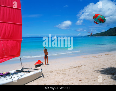 Woman standing next to catamaran on beach watching parasailing on Mahe Island in the Seychelles Stock Photo