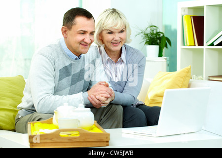 Portrait of mature man and his wife sitting in front of laptop at home and watching movie or online broadcasting Stock Photo