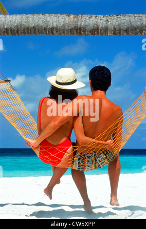 Couple sitting together in hammock under palm tree and blue skies above white sandy beach in Maldives Stock Photo