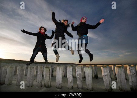 three young women jumping from wooden piles of groyne at the beach, Netherlands, Zeeland, Breskens, Sluis Stock Photo