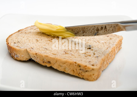Slice of granary bread being buttered on white plate against white background Stock Photo