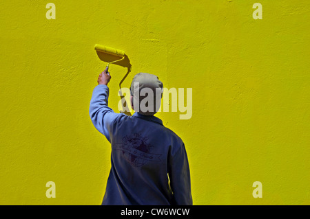 young man wearing a baseball cap is painting a house fassade yellow using a paint roller, South Africa, Bo-Kaap, Capetown Stock Photo