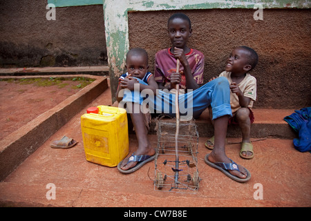 three little children with self-made toy car and water can sitting in front of house wall, Burundi, Gitega Stock Photo