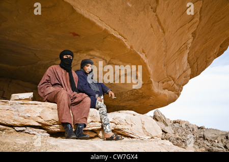 two Touaregs sitting under a rock spur at the Acacus Mountains showing a prehistoric rock painting, Libya, Sahara Stock Photo