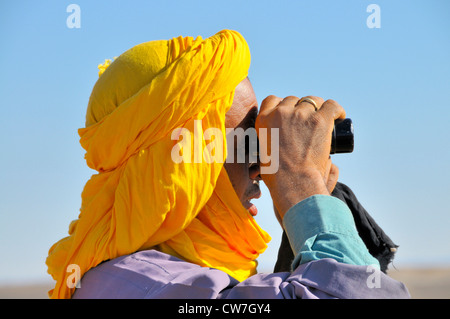 berber with traditional tagelmust searching for lost animals with a field glasses, Morocco, Erg Chebbi, Sahara Stock Photo