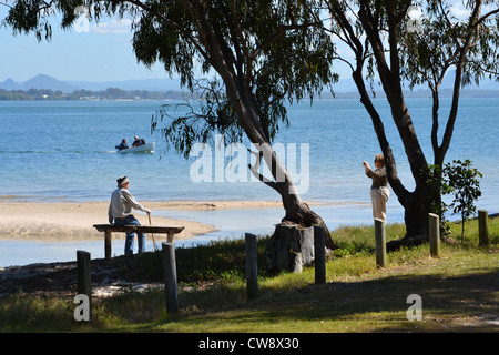 Lady taking a photo of an old man sitting on a bench on the waterfront. Bribie Island, Queensland, Australia Stock Photo