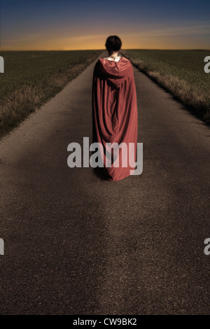 a woman in a red cape is walking along a road Stock Photo