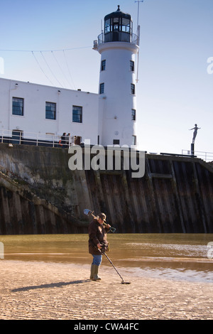 Man metal detecting on North Bay beach beneath harbour wall & lighthouse, Scarborough, North Yorkshire,UK Stock Photo