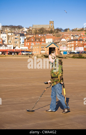 Man metal detecting on North Bay against backdrop of lifeboat station, ad old town of Scarborough, North Yorkshire, UK Stock Photo