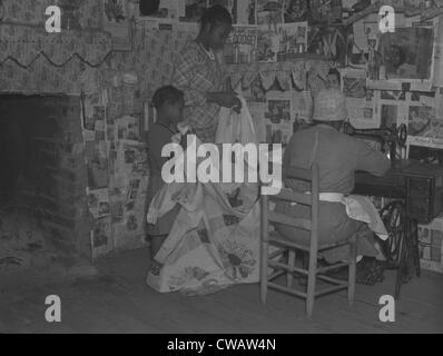 African American mother is assisted by her two daughters as she sews a quilt in their newspapered home on the Pettway Stock Photo