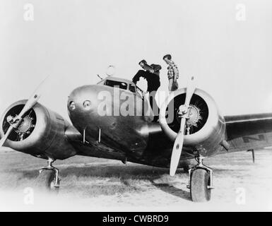 Amelia Earhart stanind on the wing of her Lockheed L-10E Electra airplane. At right is Fred Noonan, her navigator entering the Stock Photo