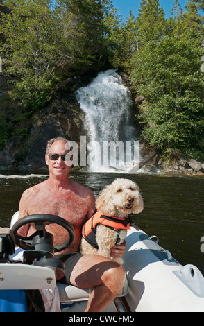 A smiling senior adult male and his cute labradoodle pet ride in a dinghy in front of Cassel Falls in Canada's Desolation Sound. Stock Photo