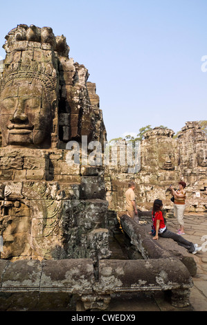 Vertical close up view of the huge carved stone faces of the Bayon temple with tourists taking photographs at Siem Reap. Stock Photo