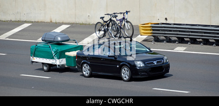 Black five door hatchback car towing collapsible camper trailer with cover & luggage box bikes on roof rack driving M25 motorway road England UK Stock Photo