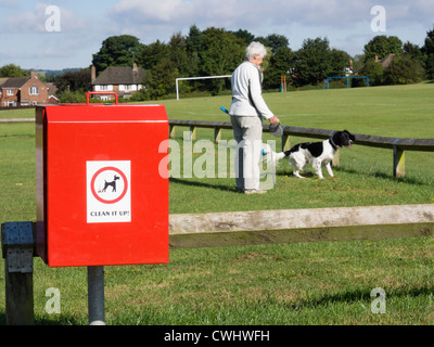 Dog waste bin by playing fields in an urban park public space with a woman walking a dog on daily walk in background. England UK Britain Stock Photo