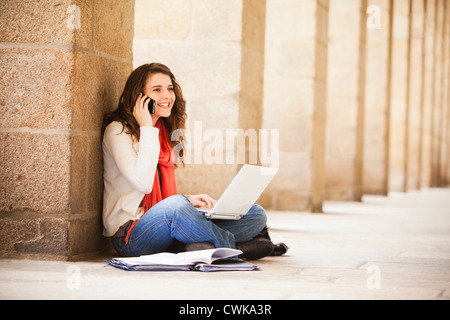 Caucasian woman sitting on ground with laptop and talking on cell phone Stock Photo
