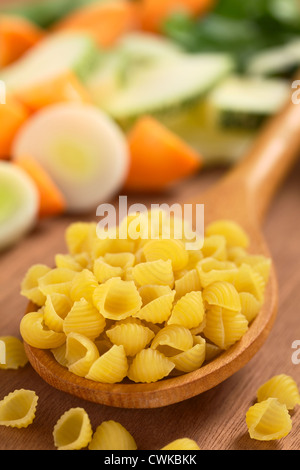 Raw shell pasta or conchiglioni on wooden spoon with raw vegetables (carrot, leek, zucchini) in the back Stock Photo