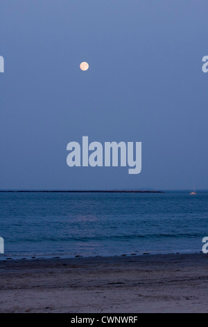 Shoreline at Dusk. High Tide on the Atlantic Ocean as a Full Moon rises over a jetty where a sailboat is protected at anchor Stock Photo