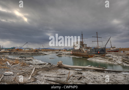 A shipwreck at a gloomy scrapyard in Alaska where the sky is grey and the ground is covered with the dust of a recent volcano. Stock Photo