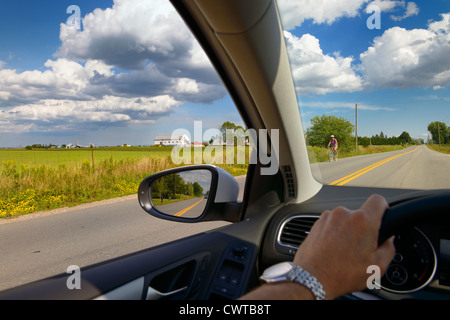 Hand of man on steering wheel of car driving down a country road with farm bicyclist and clouds Stock Photo