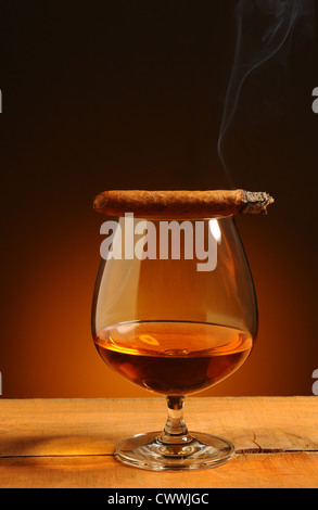 Brandy glass with a lit cigar on its rim over a warm background. Smoke trail wafting up from cigar. Stock Photo