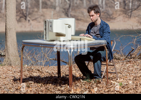 A Young Man typing on his Computer in the Outdoors and surrounded by trees, mother nature and a beautiful blue sky. Stock Photo