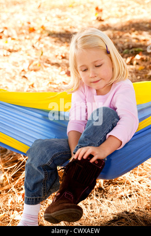 A little blond girl sitting on a hammock putting on her boots. Stock Photo