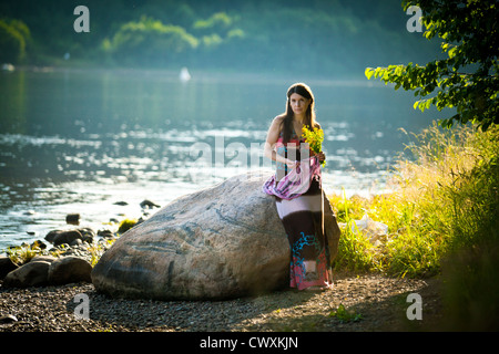 Portrait of a beautiful young woman stands near the stone on the bank of the river Stock Photo