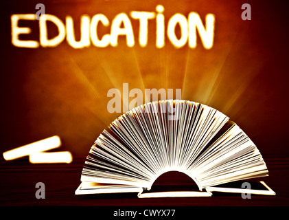Image of open book on wooden school desk in classroom, textbook over brown chalkboard, magic book with glowing light Stock Photo