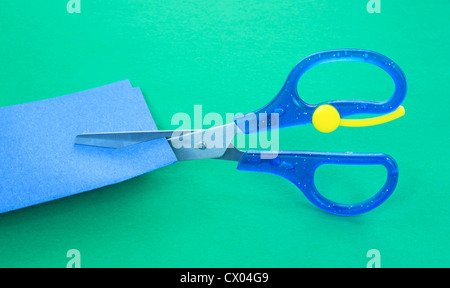 Child's scissors cutting blue craft paper on a green paper background. Stock Photo