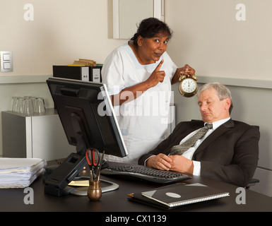 Cleaning woman holding an alarm clock next to a sleeping office worker Stock Photo