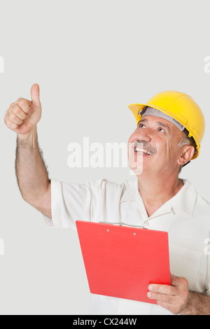 Happy senior constructor holding clipboard while gesturing thumbs up over gray background Stock Photo